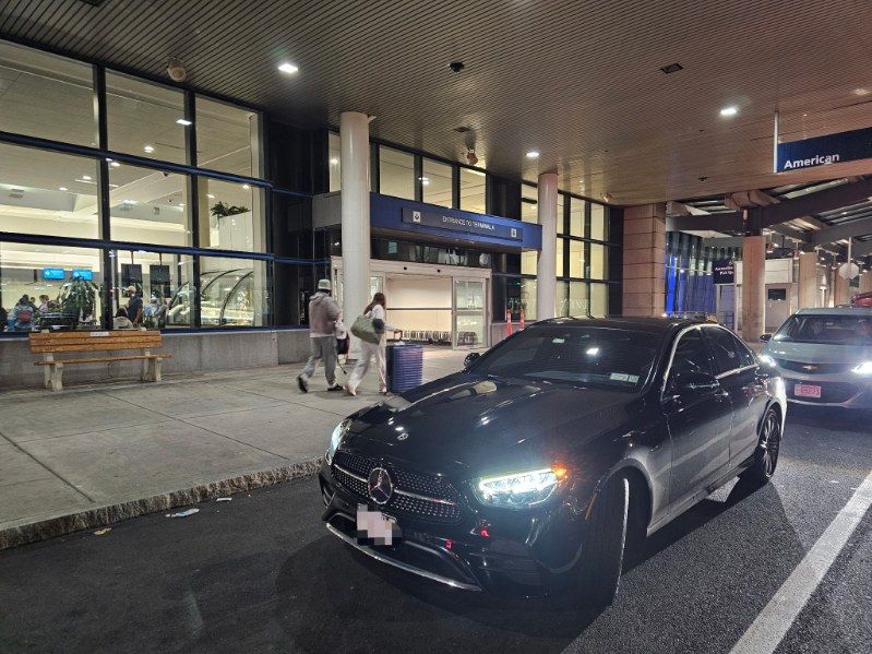 Black car parked at airport entrance; people walking near building with glass windows, bench visible.