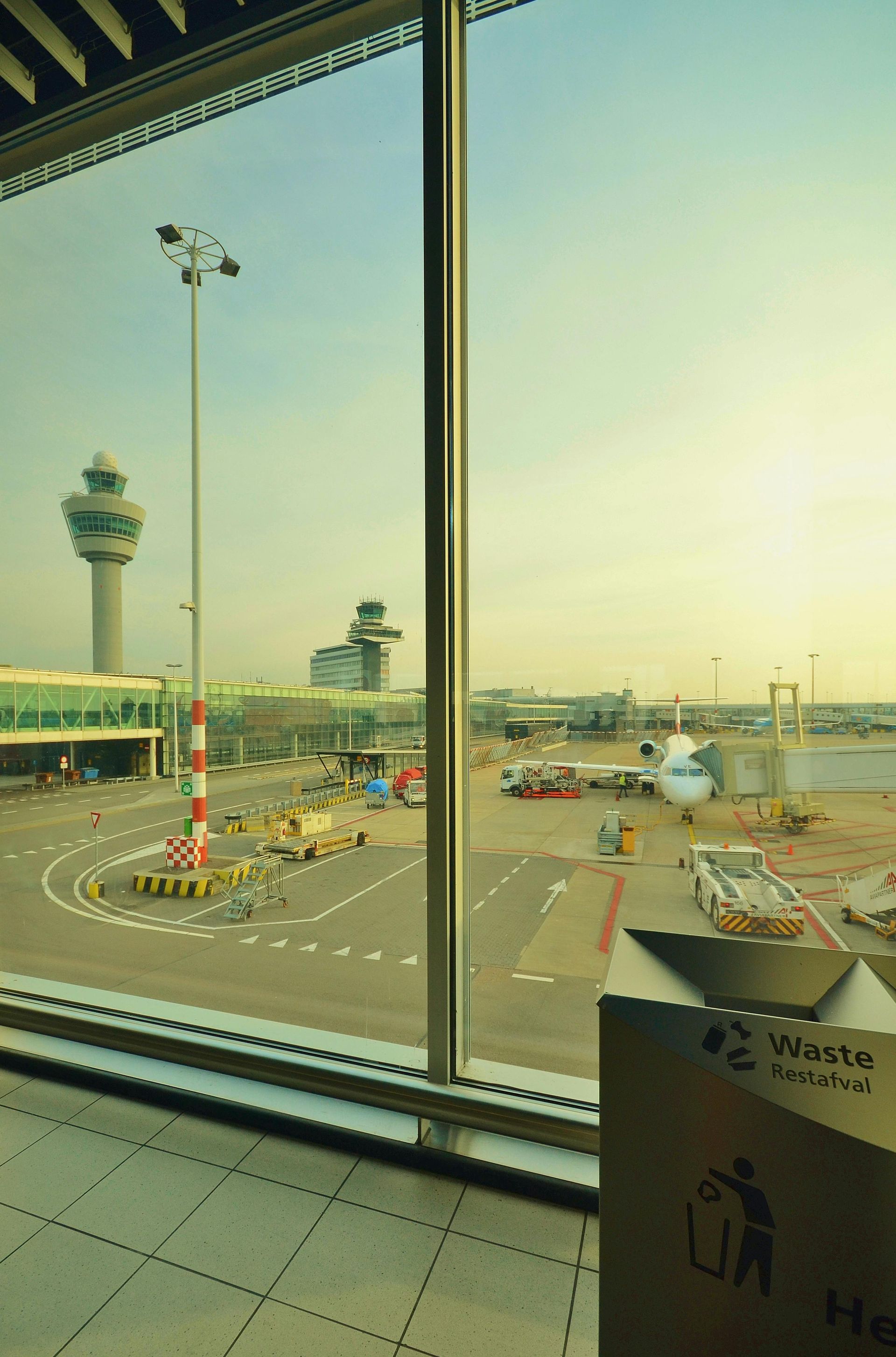 Airport view through a window, including control tower, runway, parked airplanes, and a trash can.