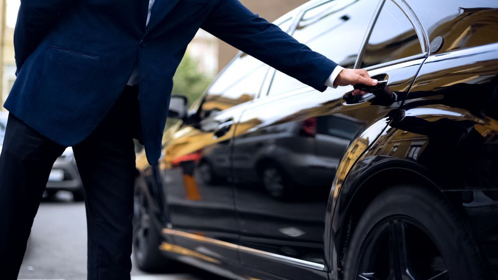 Man in suit opening a black car door on a city street.