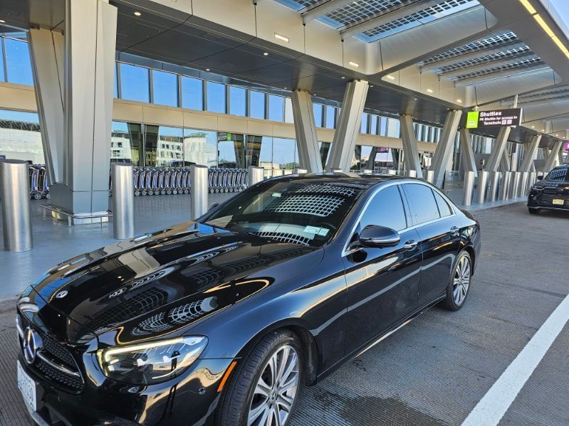 Black Mercedes-Benz sedan at an airport curb, likely for pickup. Reflective glass windows, sunny day.