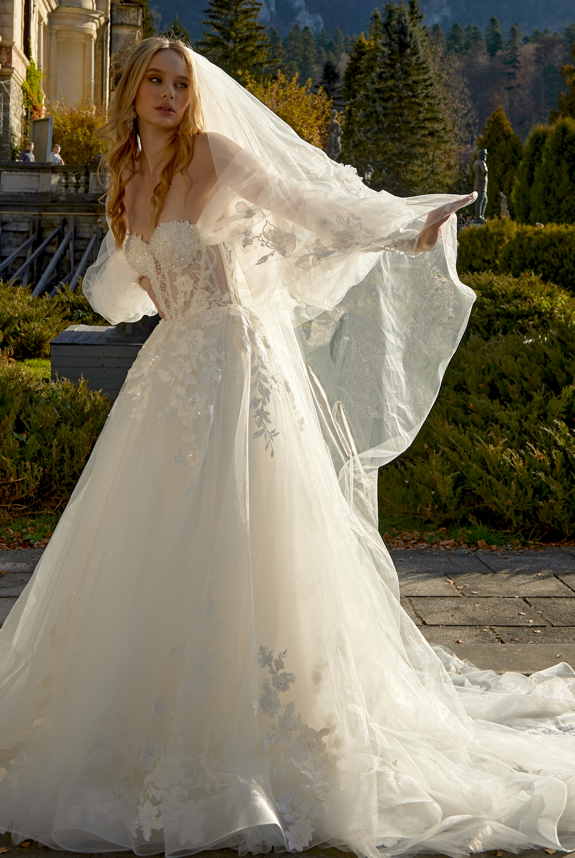 Bride in a white wedding gown standing outdoors by a garden with mountains and trees in the background