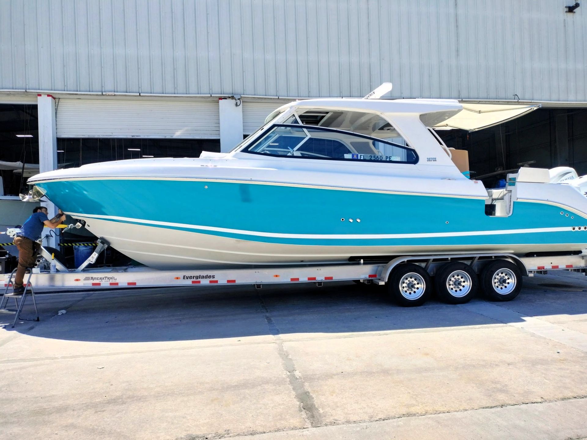A teal and white boat on a trailer, parked in front of a building. A person is nearby.