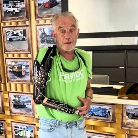 Man with a prosthetic arm stands indoors wearing a green shirt and jeans. Wall of framed photos behind him.