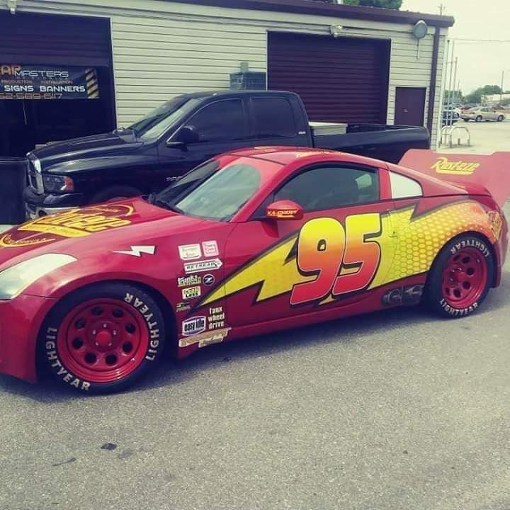 Red Lightning McQueen car wrap on a Nissan sports car parked near a garage. Black truck in background.