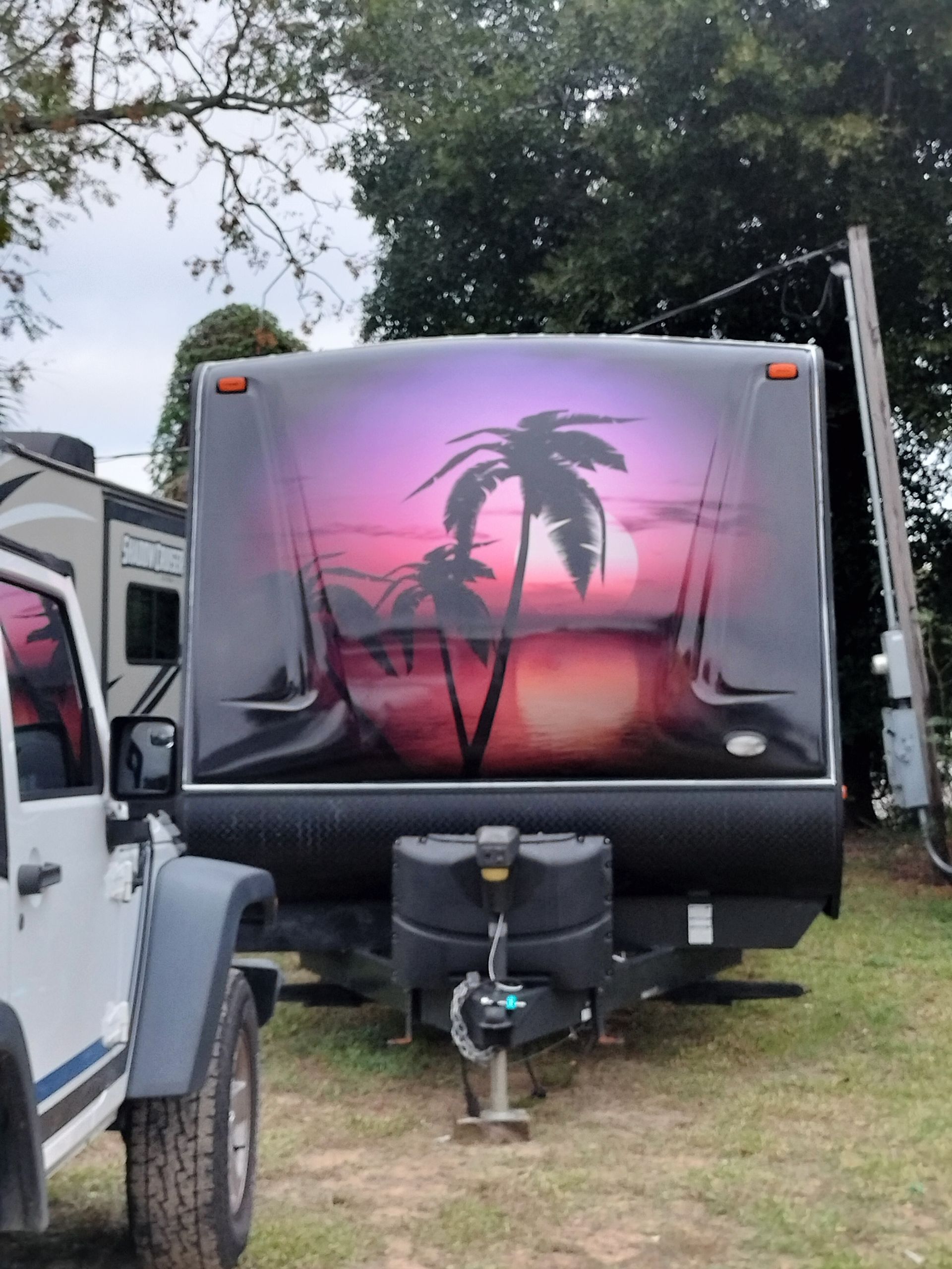 Black travel trailer with a sunset scene painted on its front. A Jeep is to the left.