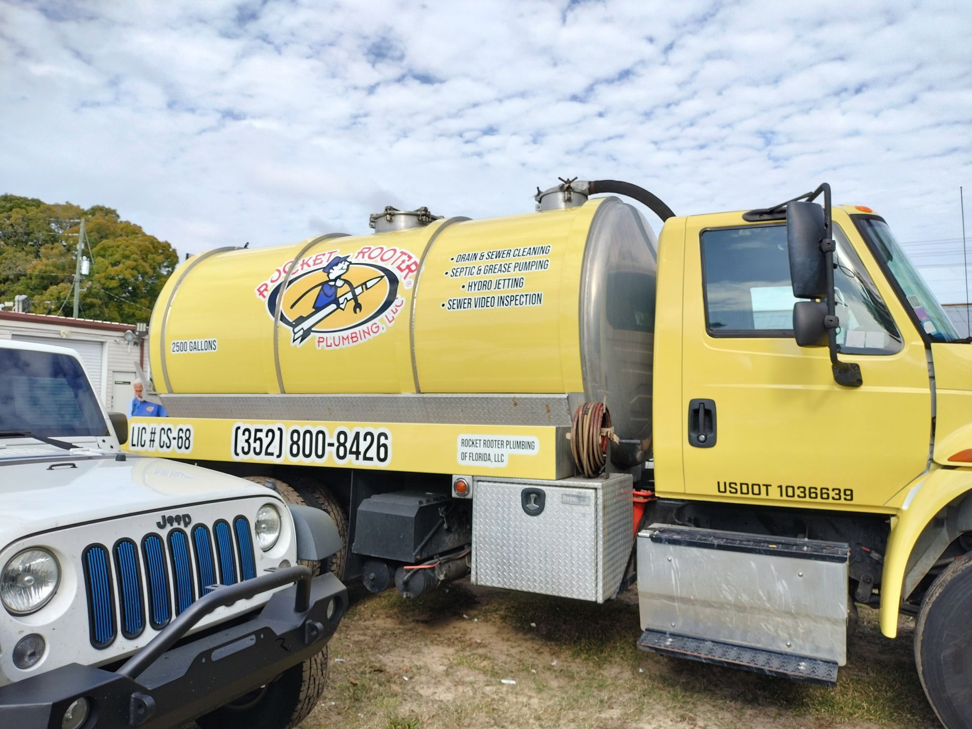 Yellow septic tank truck next to a white Jeep on a cloudy day.