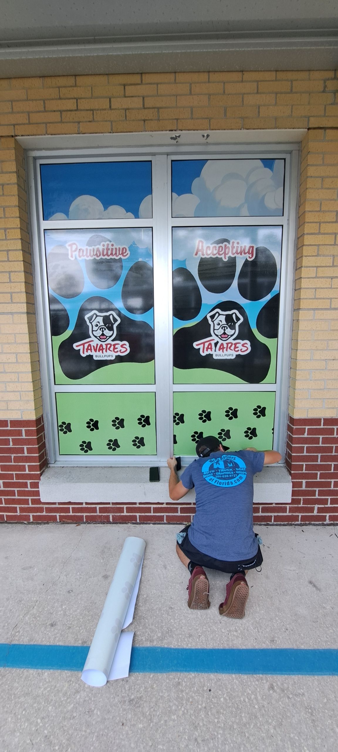 Person kneeling at a storefront window, installing decals. Storefront has dog paw graphics. Rolls of material on the ground.