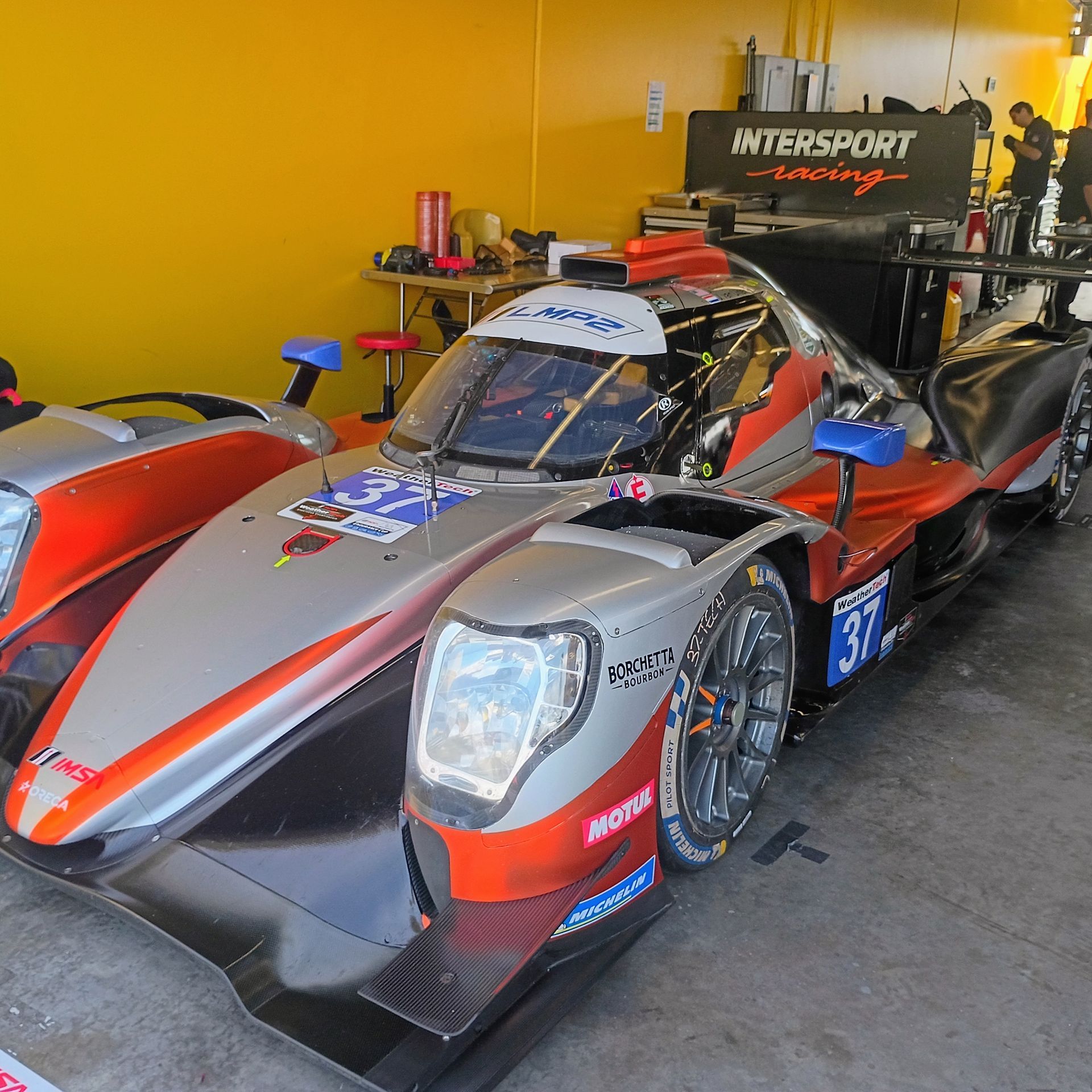 Silver and orange race car in a garage with “Intersport Racing” signage.