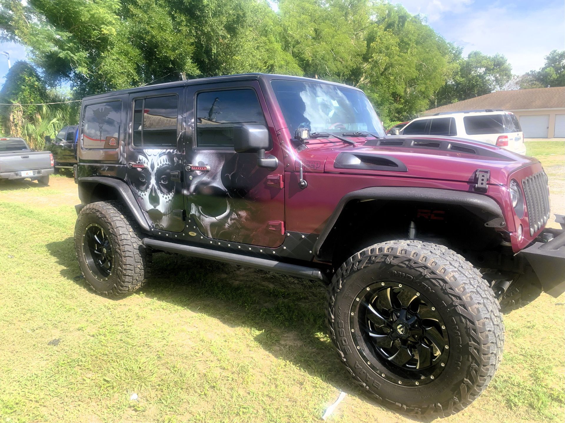 Maroon Jeep Wrangler with skull graphics and black wheels parked on grass.