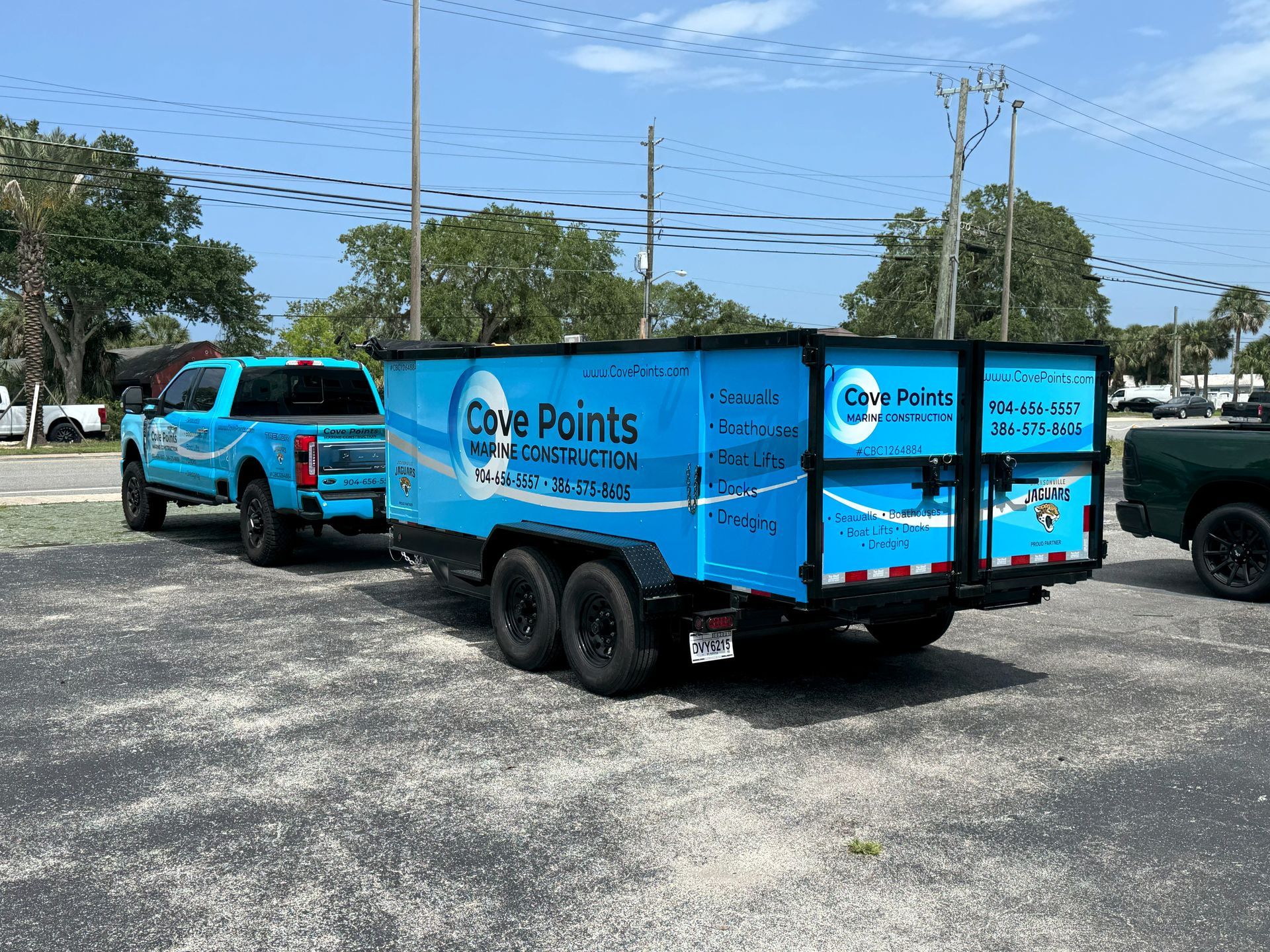 Blue truck towing a blue trailer with company logos parked on a gravel lot under a sunny sky.