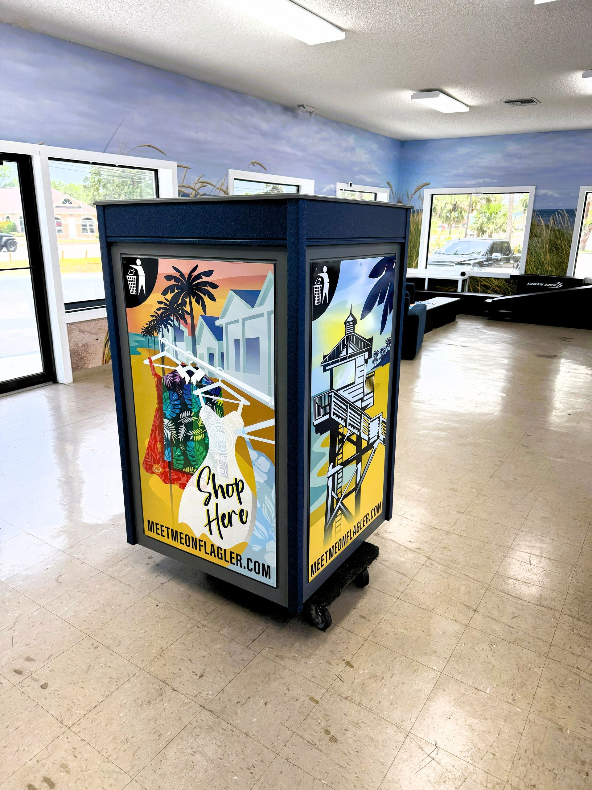 A rolling display with colorful tropical artwork stands inside a room with large windows, light flooring, and blue walls.