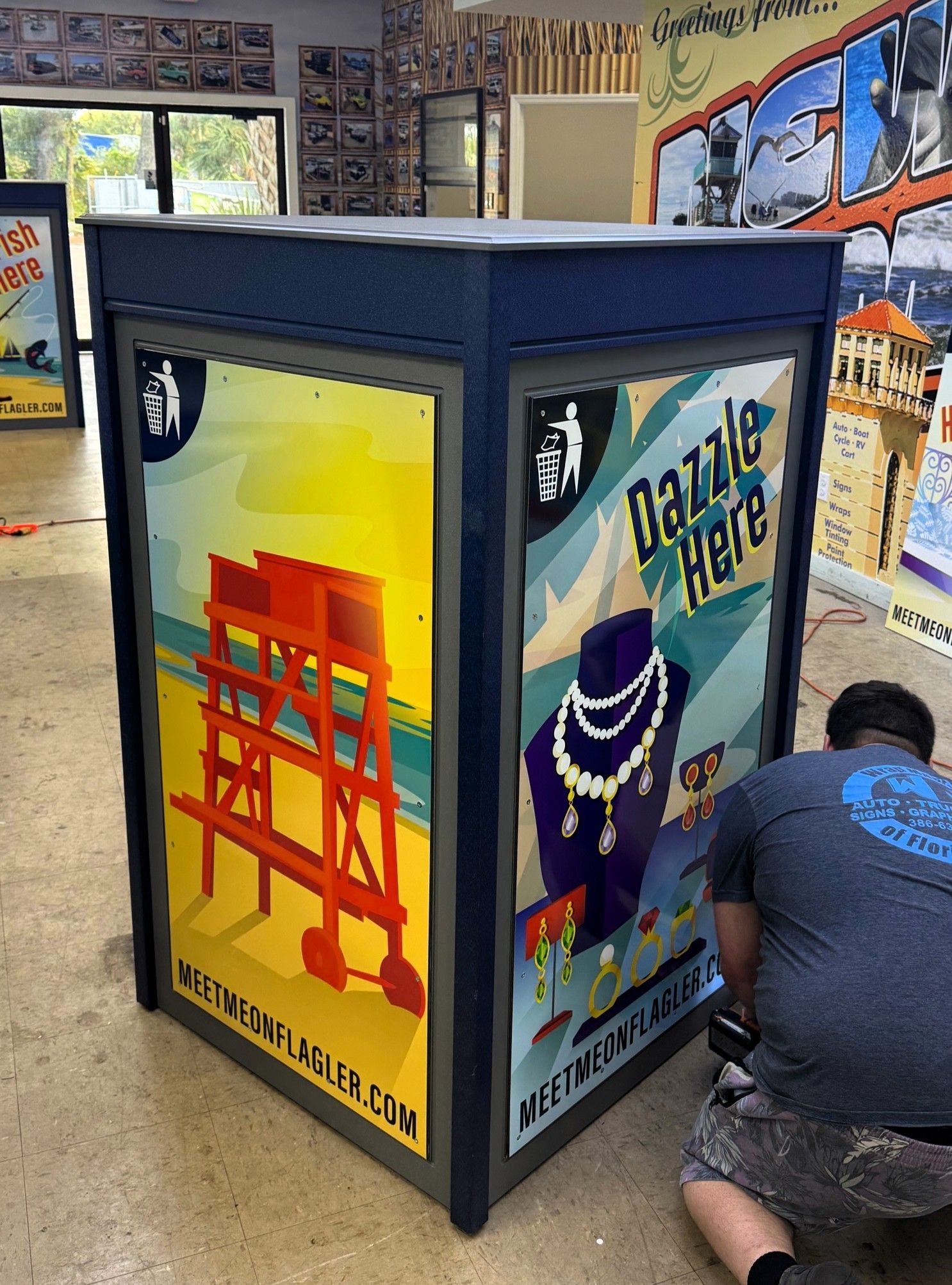 A person works on a large, multi-sided kiosk with posters of a lifeguard tower and jewelry, in a room.