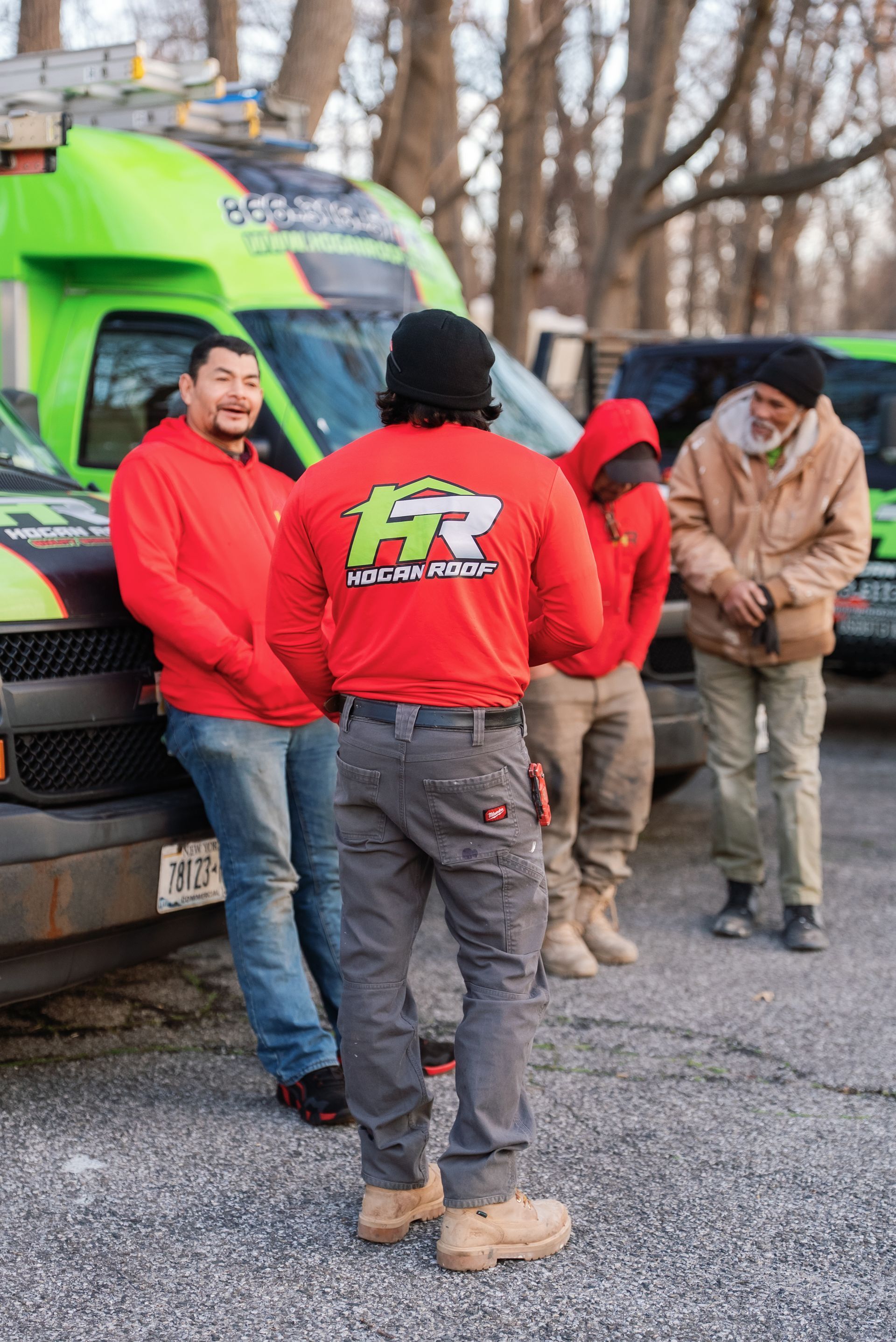 Three workers in red jackets stand beside a green truck on a gravel lot, talking outdoors.