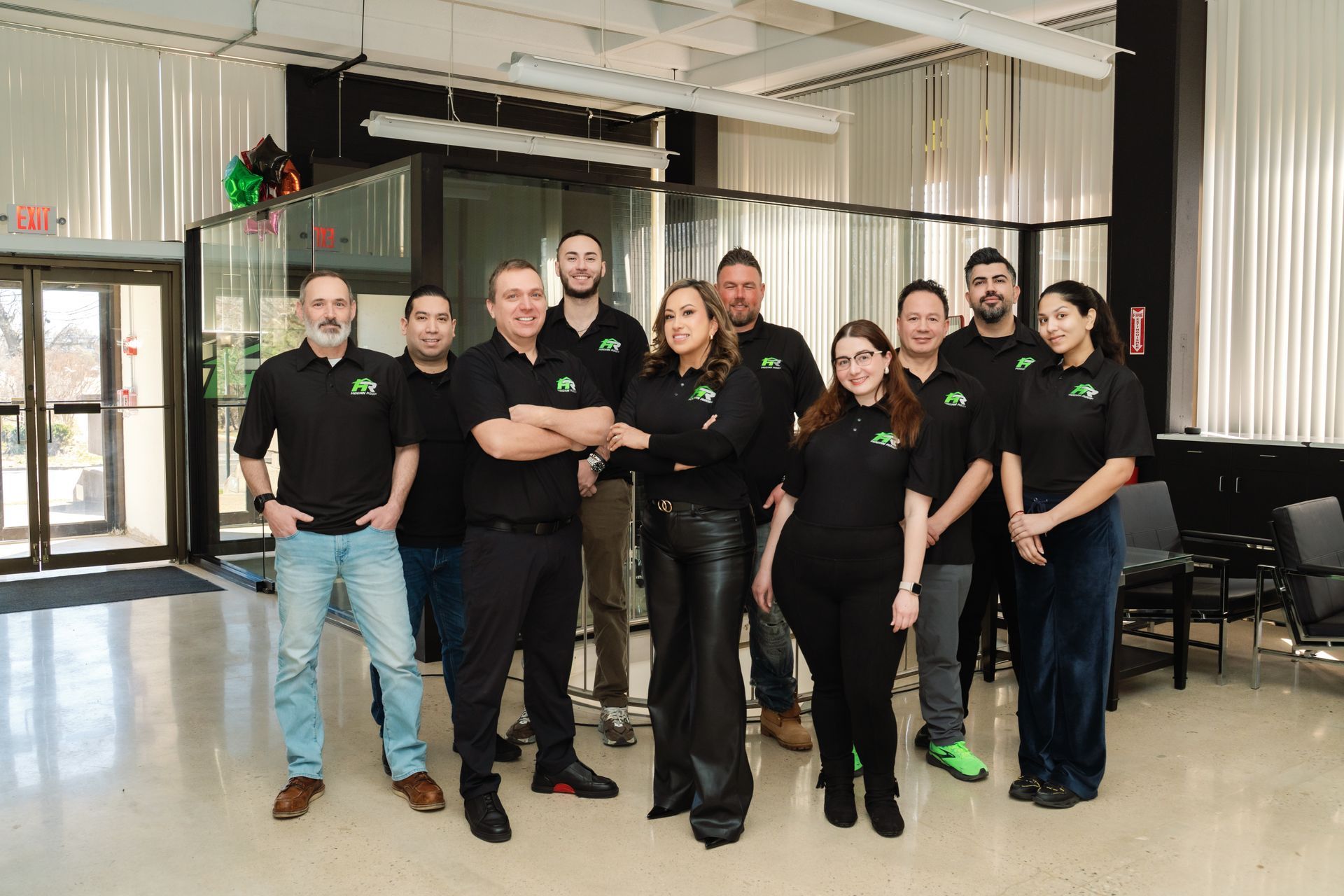 Group of people in black shirts posing in an office lobby, with glass walls and bright lights.