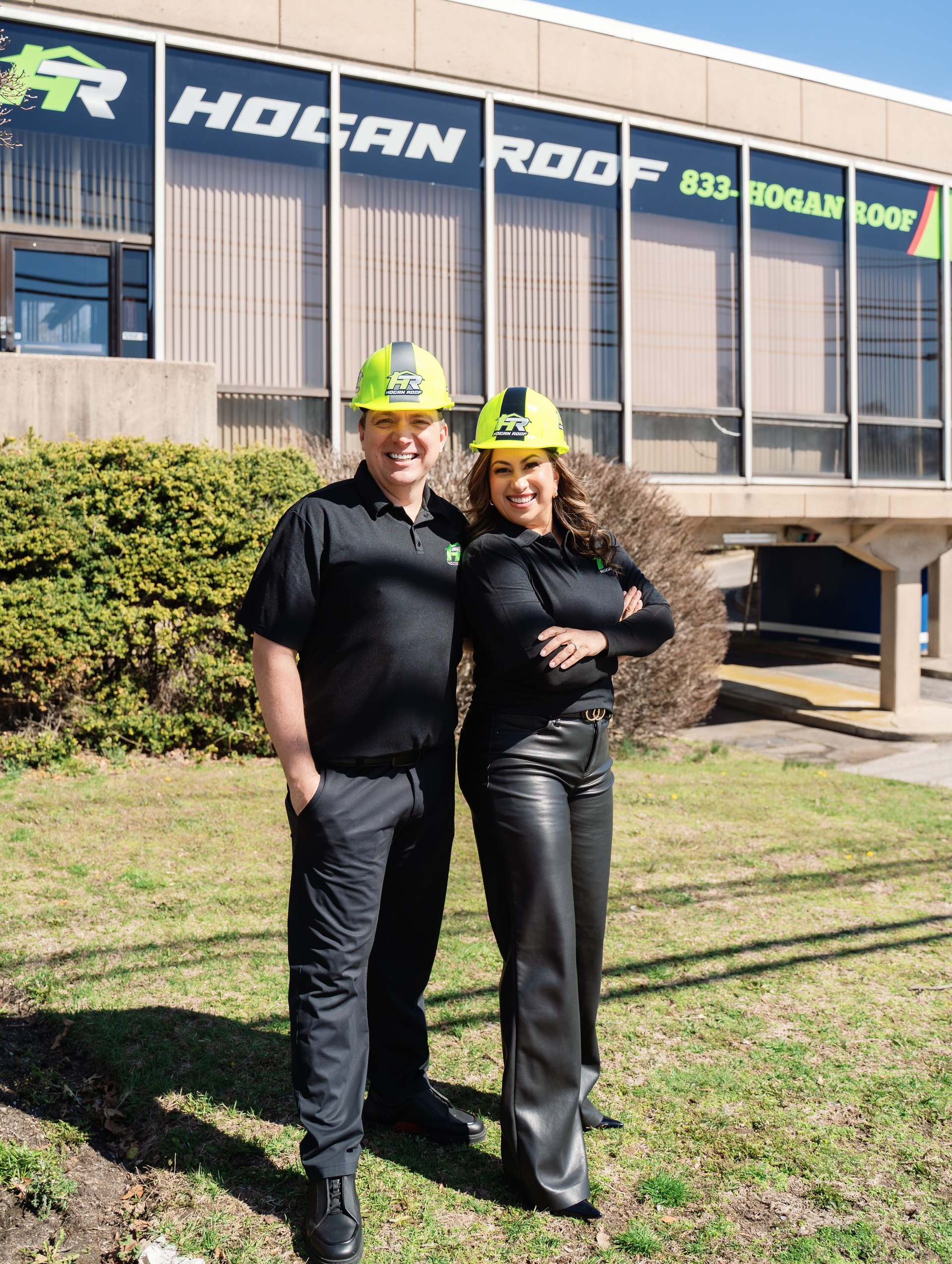 Two people in hard hats and black clothing stand smiling outside a building with a large sign.