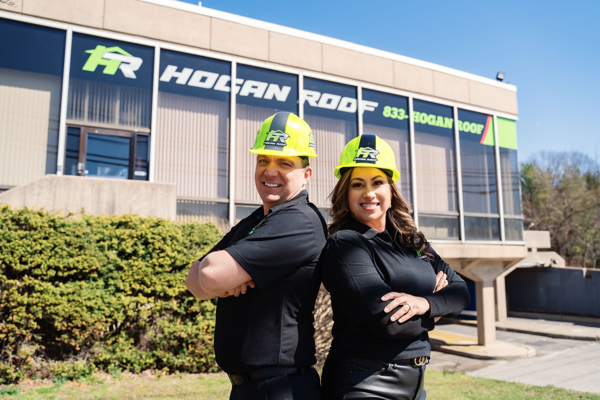 Two workers in yellow hard hats standing back-to-back outside the Hogan Roads building, smiling.