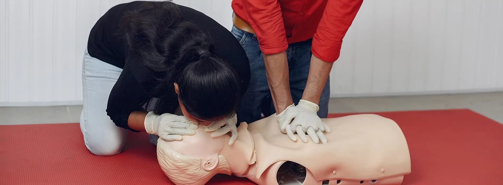 Two people practice CPR on a dummy. One kneels by the head, the other performs chest compressions.