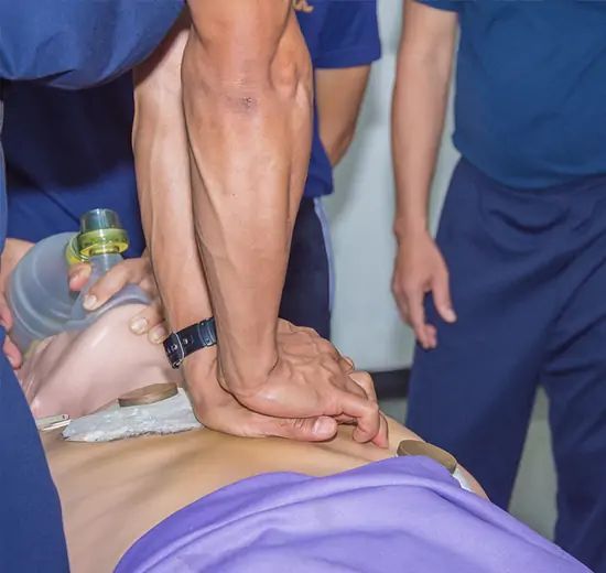 Hands performing CPR on a training mannequin while another person provides ventilation with a bag valve mask.