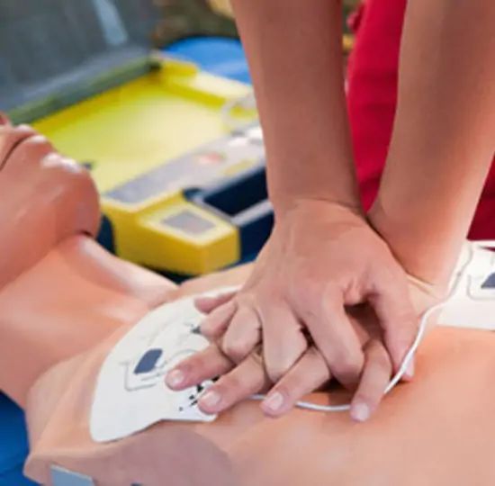 Person performing chest compressions on a CPR training mannequin with an AED nearby.