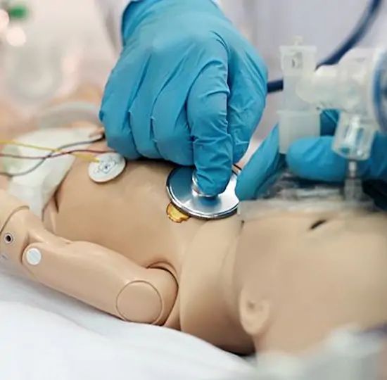 Hands in blue gloves using a stethoscope on a baby-sized medical training mannequin.