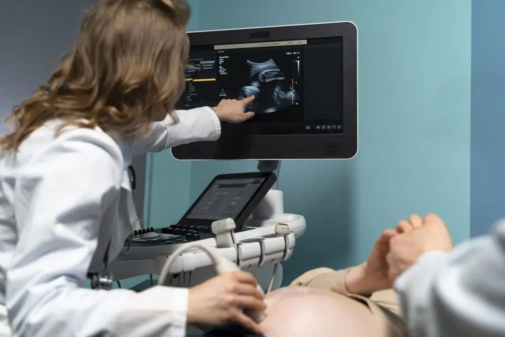 Doctor points at ultrasound scan on a screen. Patient lies on a bed, receiving the scan.