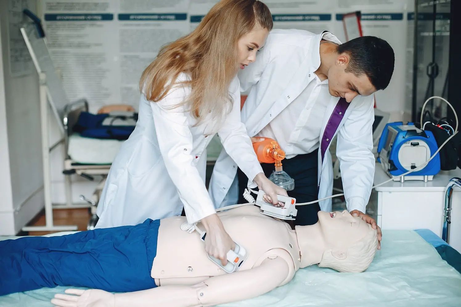 Two people in white lab coats practice using a defibrillator on a dummy in a medical setting.