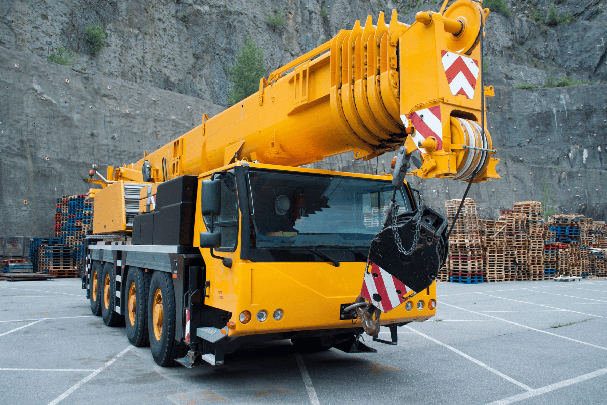 Yellow mobile crane truck parked in a quarry, with stacked pallets in the background.