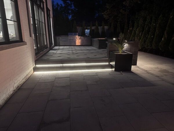 A modern outdoor patio at night with stone pavers, illuminated steps, and a lit planter beside a house exterior.