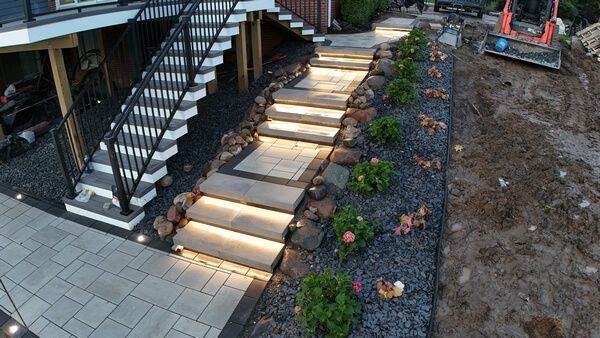 Illuminated stone steps leading outdoors next to a dark staircase and a landscape bed with gravel and small plants.