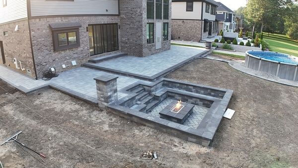 A sunken stone fire pit area with built-in seating, steps, and a lit fire feature on a gray patio next to a house.
