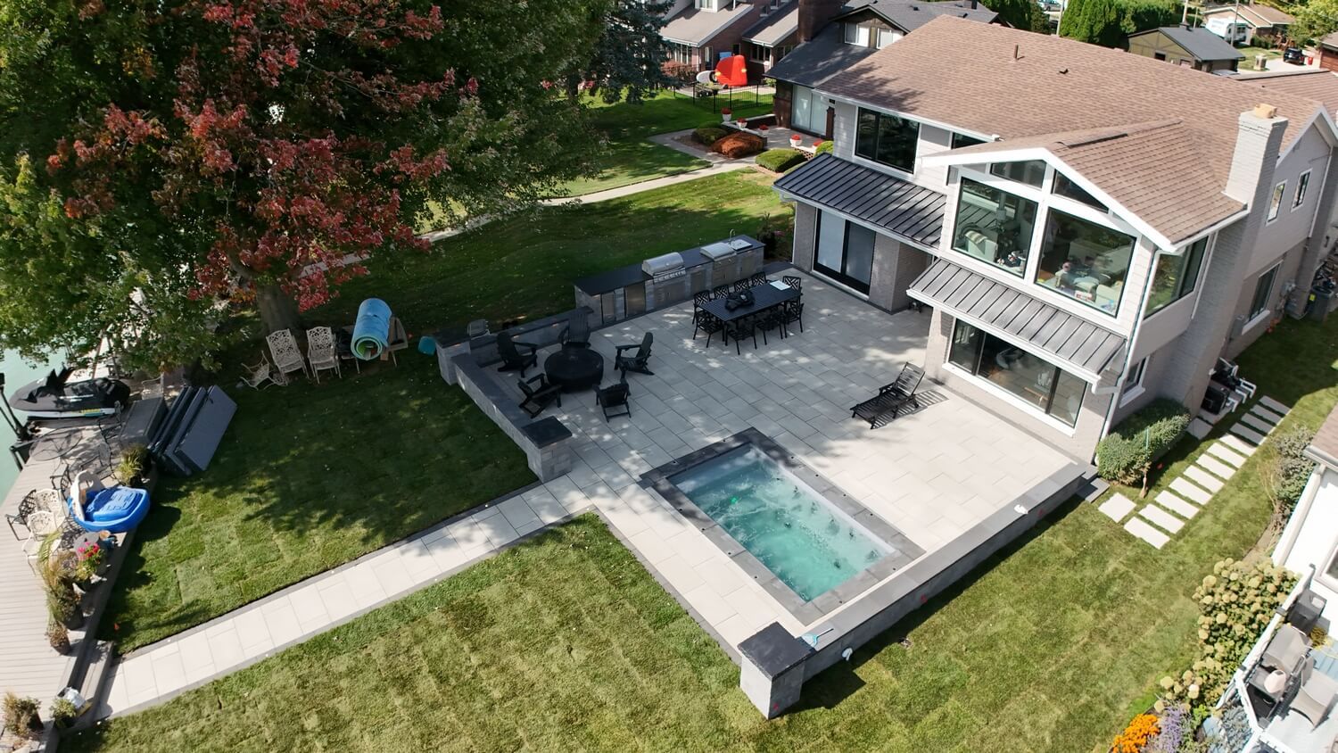An aerial view of a two-story house with a backyard patio featuring an in-ground pool, dining area, and lawn near a lake.