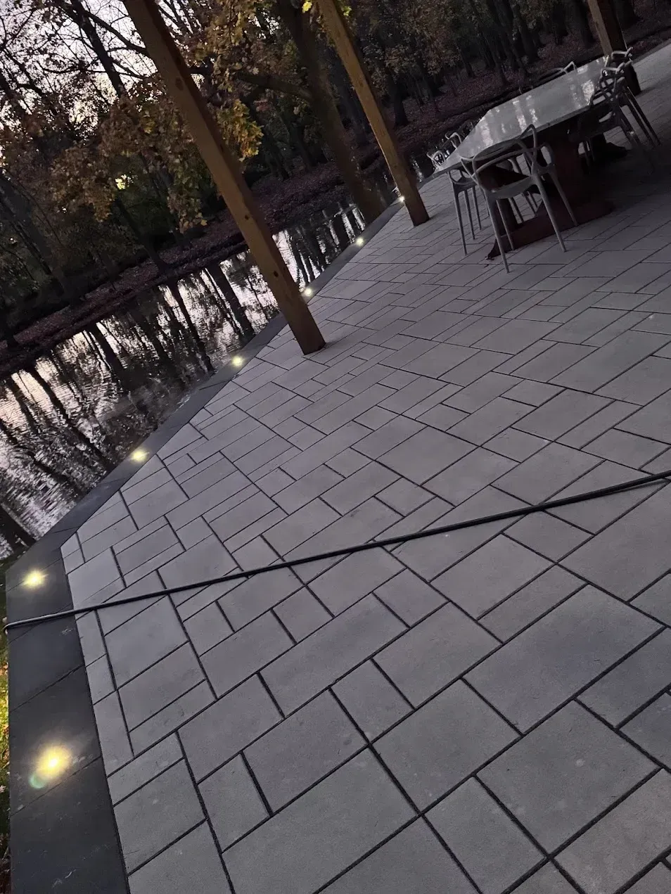 A patio with gray rectangular pavers and inset circular lights, bordered by a dark edge near a lake at dusk.