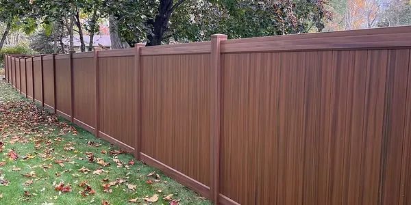 A long, brown privacy fence with vertical panels stands in a grass lawn covered with scattered autumn leaves.