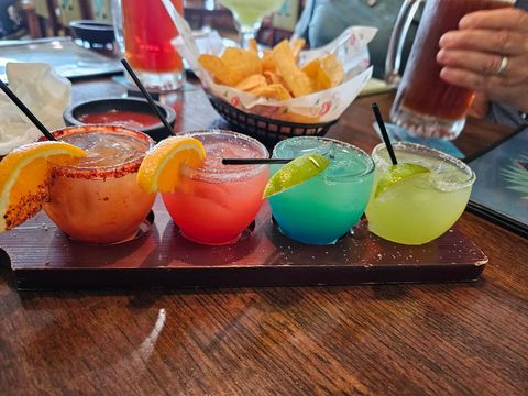 Four colorful margarita shots with orange slices on a wooden table, with chips and salsa in the background