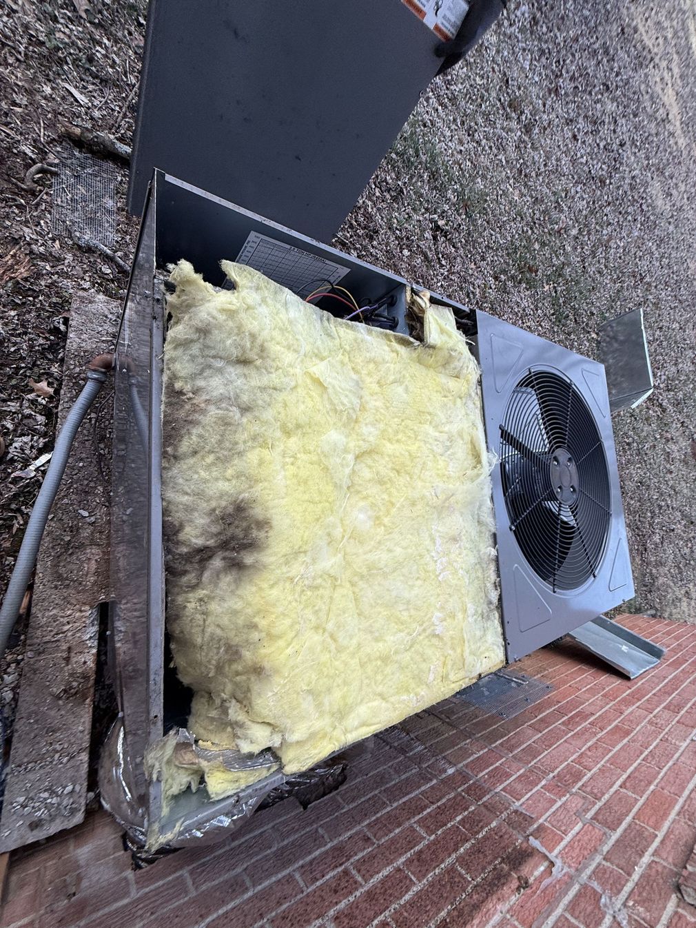 Damaged rooftop HVAC unit with exposed yellow insulation beside a brick wall and gravel surface