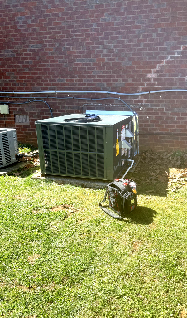 Outdoor HVAC unit beside a brick wall on a grassy lawn, with a small plant in front.
