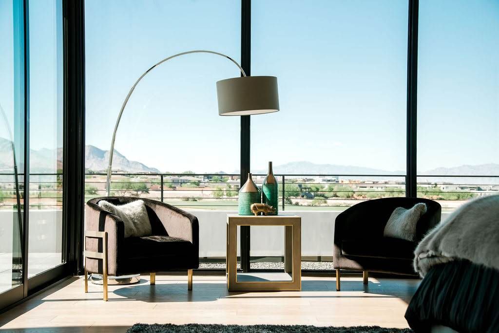 Two dark velvet armchairs facing a mountain view, under a large arc lamp, next to a small wooden table with decorative bottles.