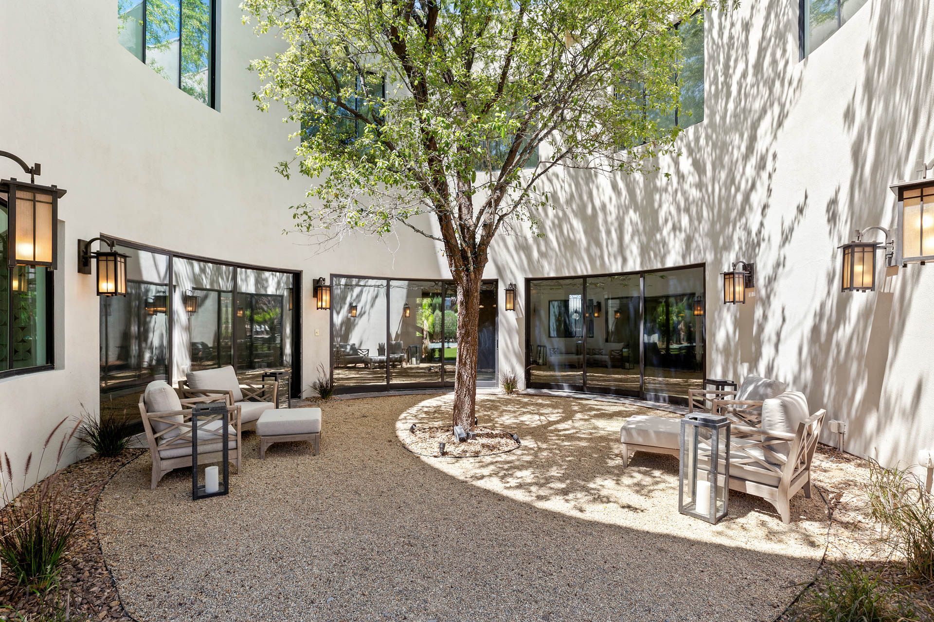 Courtyard with tree, seating, and windows against white walls. Gravel ground, sconces. Sunny.