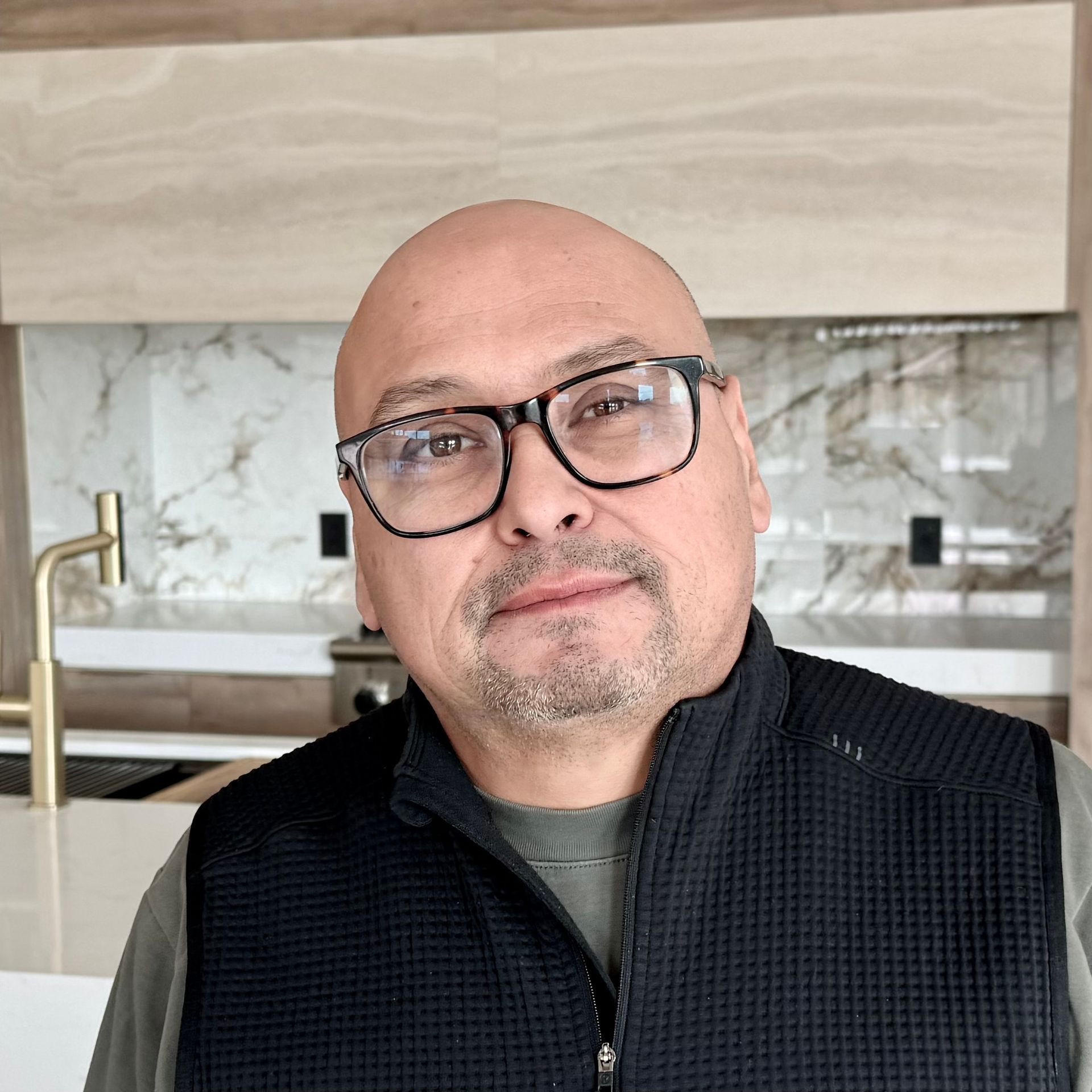 Man wearing glasses, black vest, and neutral-colored shirt, set in a modern kitchen.