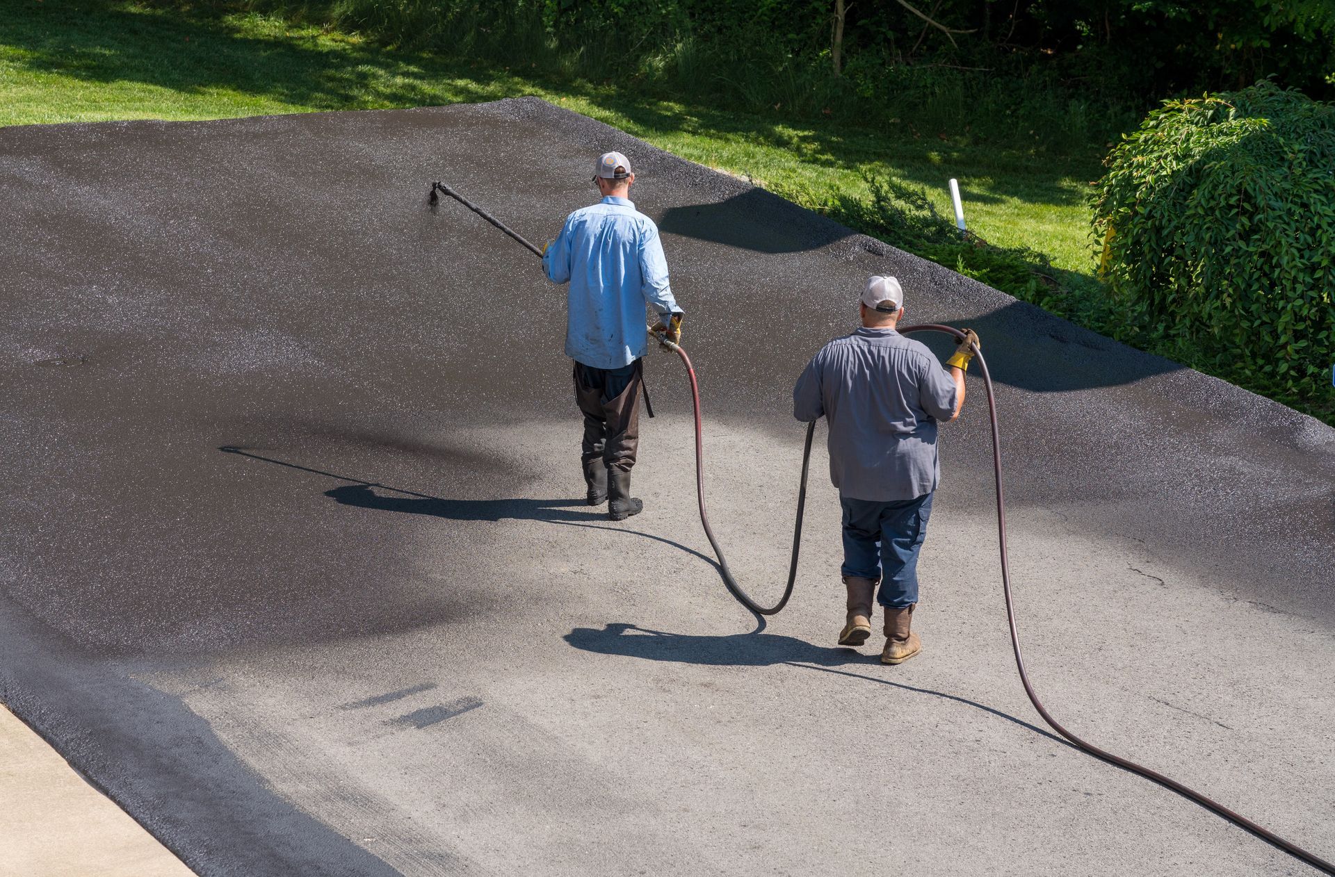 Two workers in casual clothing use a spray hose to apply a dark sealant to an asphalt driveway.