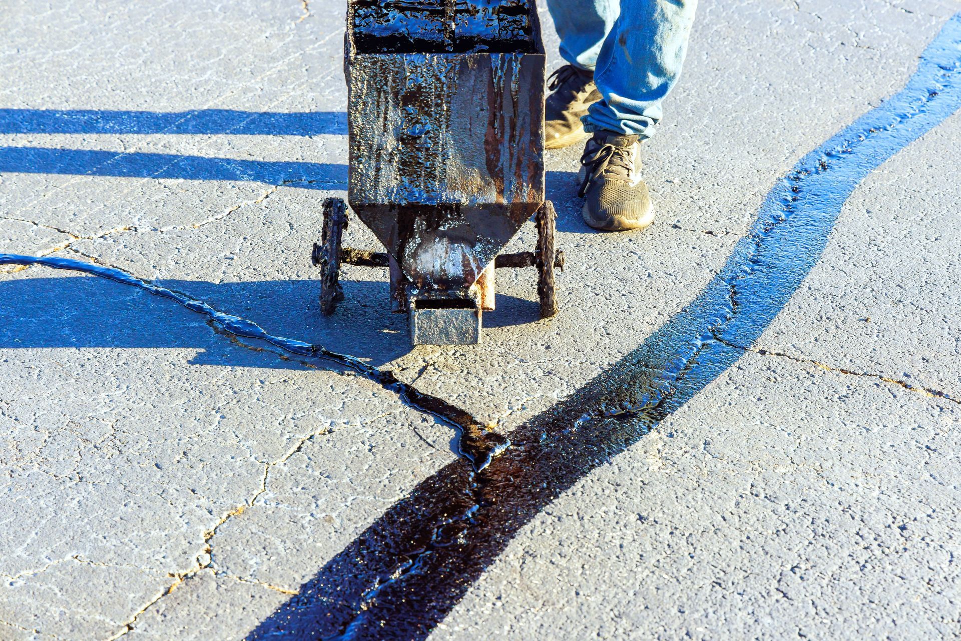 A person uses a push machine to apply hot, black liquid sealant into a crack on a grey asphalt surface.
