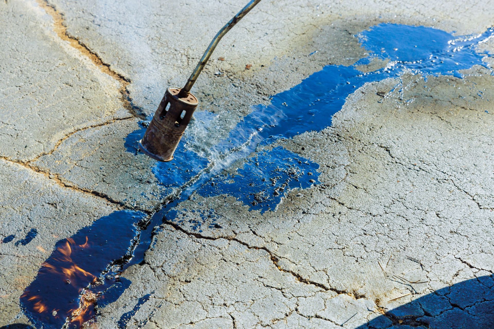 A propane torch melts black sealant into a cracked concrete surface to repair the pavement.