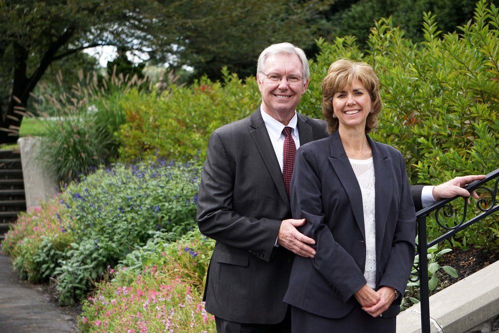 Smiling couple in business attire standing by a garden walkway with greenery and flowers