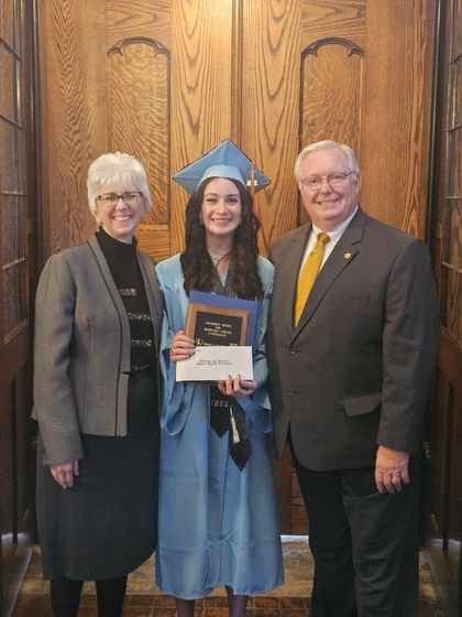 Graduate in cap and gown holding diploma, standing between two people in front of wooden doors.