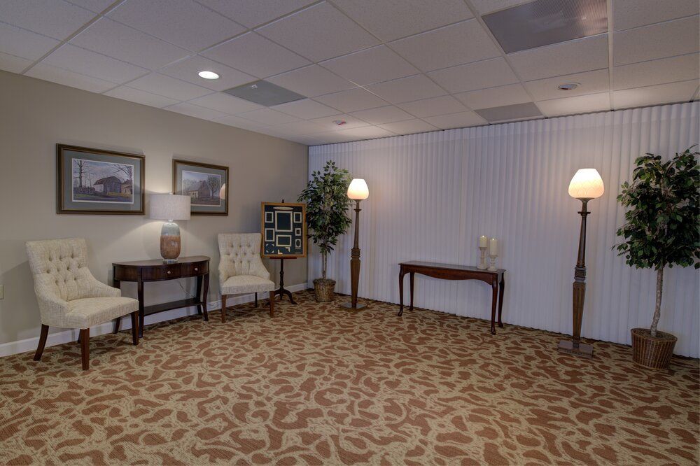 Quiet lobby seating area with two chairs, table, lamp, and potted plants on patterned carpet.