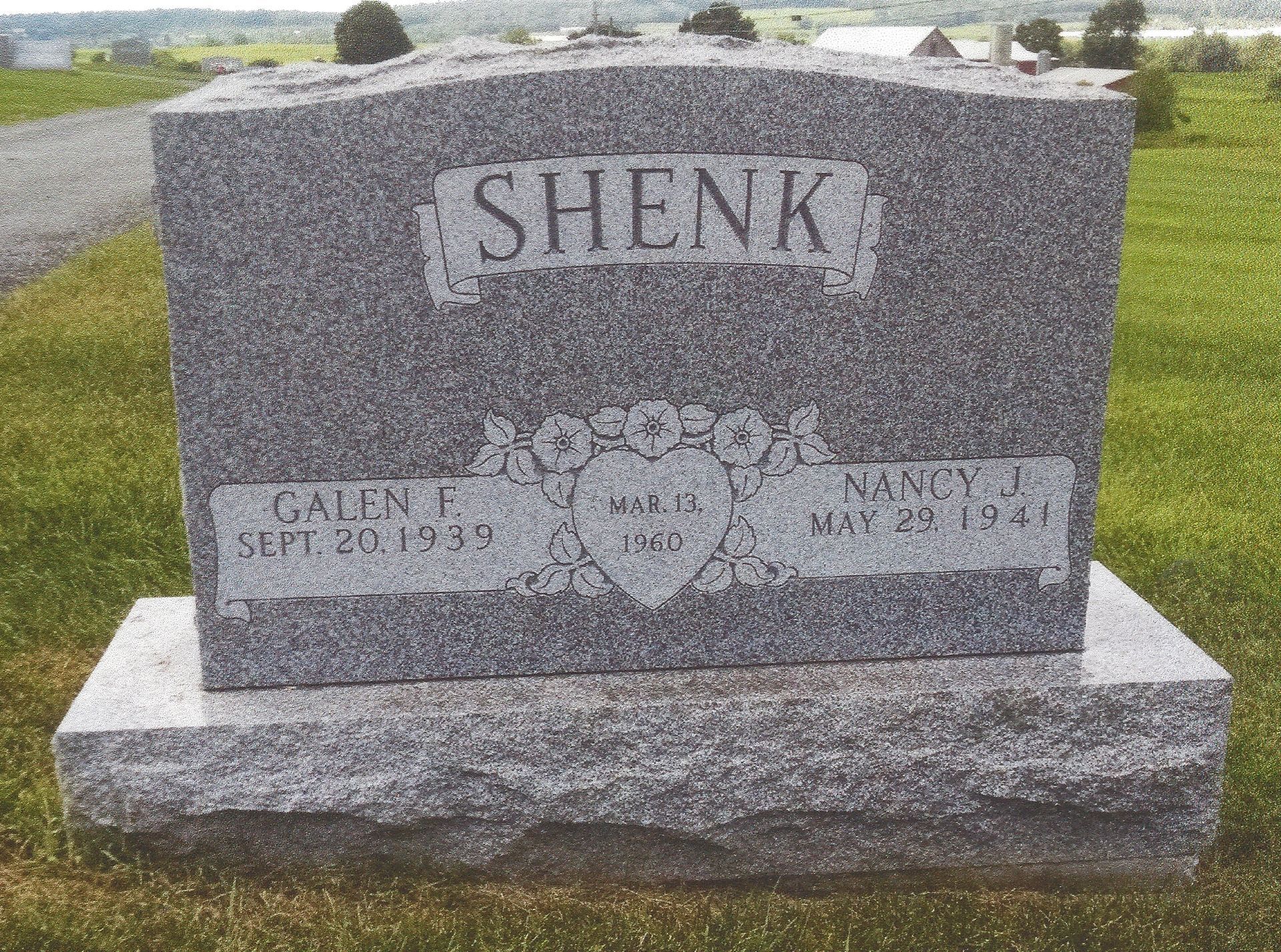 Gray gravestone engraved “SHENK” in a cemetery with grass and other headstones in the background