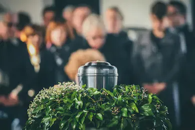 A silver urn surrounded by green foliage at a memorial, with blurred mourners in the background