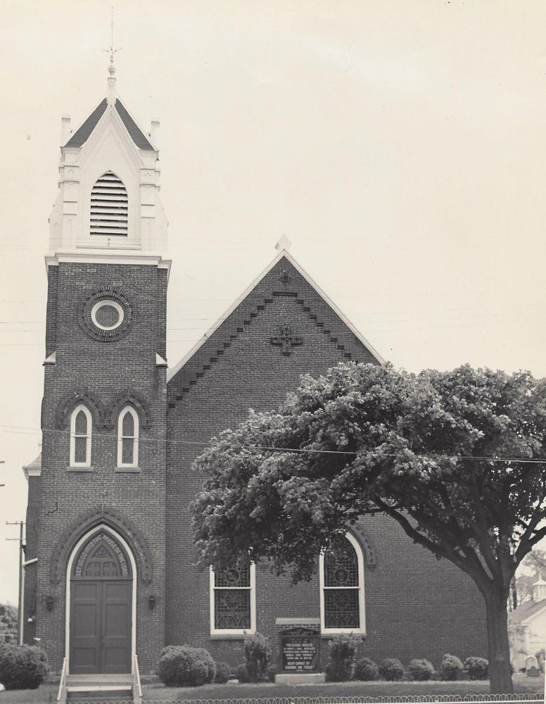 Brick church with tall steeple and arched windows beside a large tree