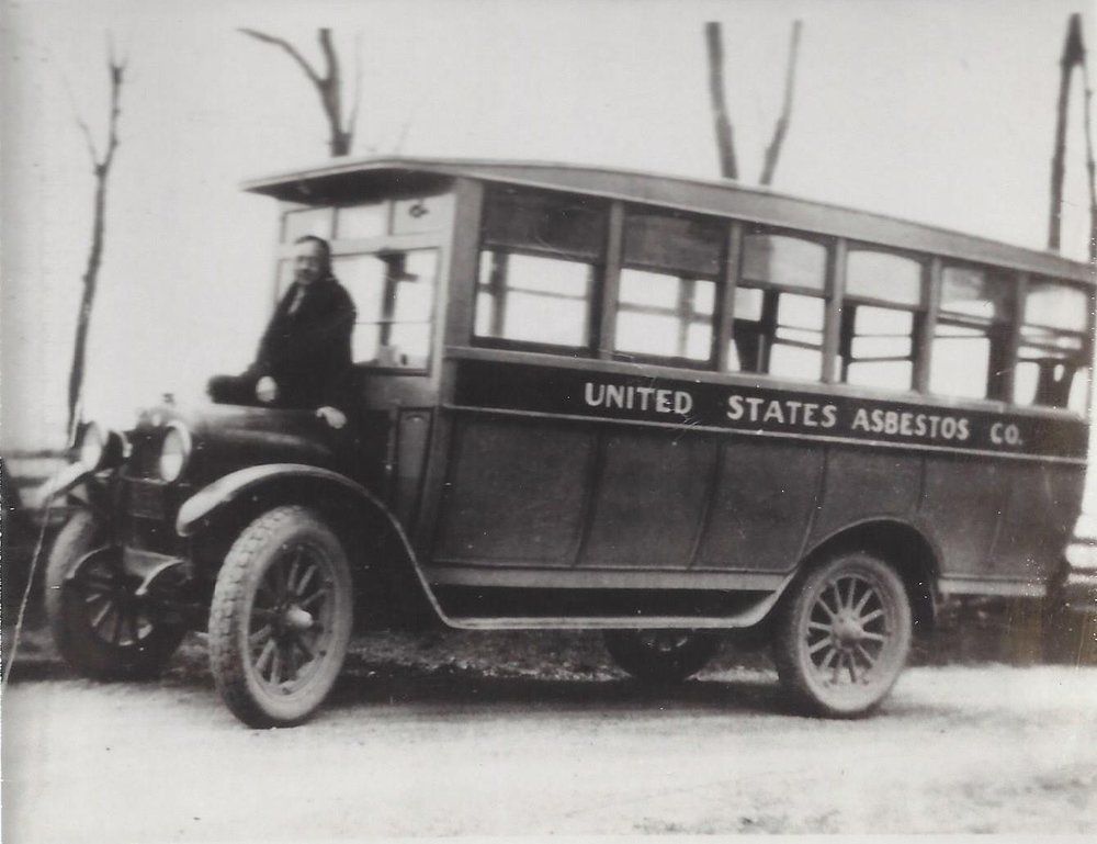Black-and-white photo of a vintage bus labeled “United States Asbestos Co.” with a driver beside it.