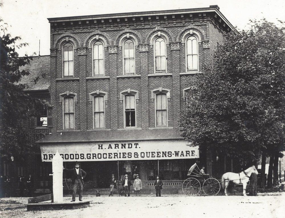 Historic black-and-white storefront of H. Arndt, a groceries and queensware shop, with people and bicycles outside.